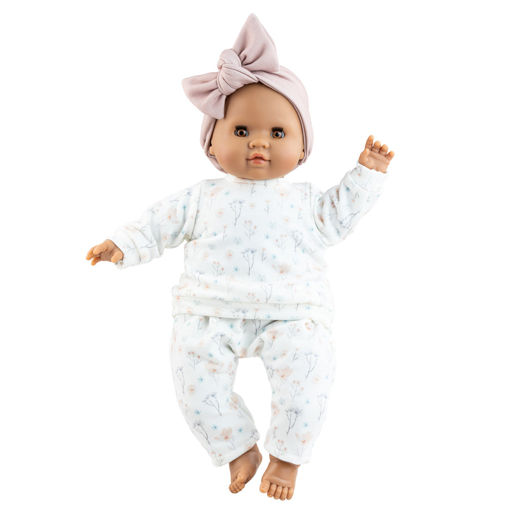 Die Puppe Alberta trägt ein schlichtes Puppenoutfit mit zarten Blumen und einer fliederfarbenen Schleife. Die Puppe hat eine dunklere Hautfarbe und braune Augen. 