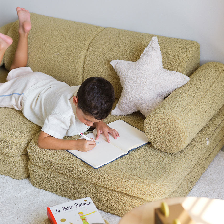 Junge liegt auf dem Bauch auf dem Kindersofa von Wigiwama und schreibt in einem Heft. Perfekt zum Erinnerungen sammeln. 
